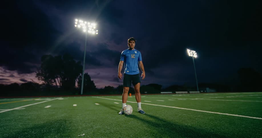 Lights illuminate the soccer field as a player stands with a soccer ball at his feet