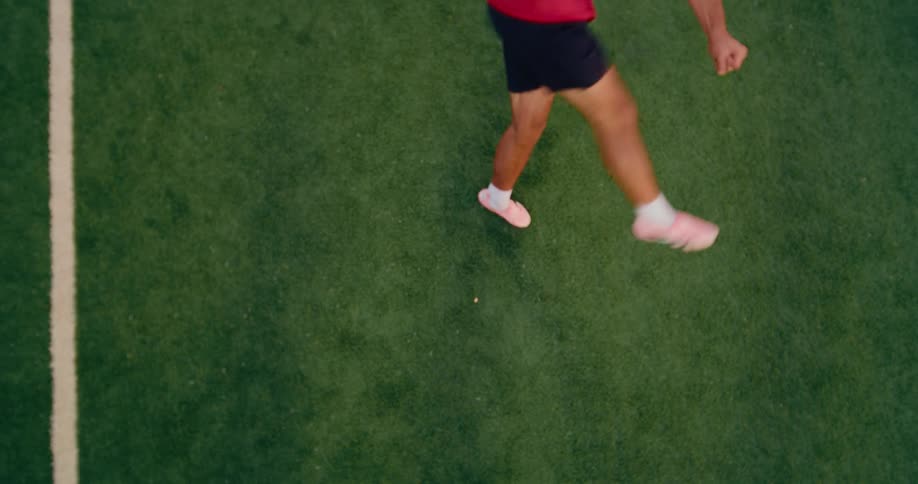 A topdown view of an athlete running across a green turf field with a white sideline a soccer ball rolls across the frame as the persons legs and pink shoes move quickly past