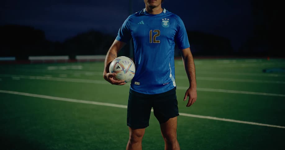 A man stands on a soccer pitch holding a soccer ball at night