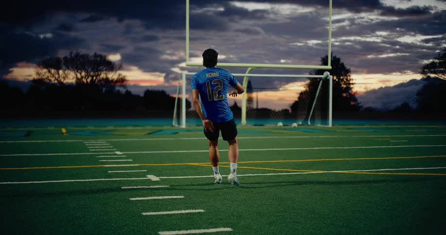Young adult man wearing sports uniform kicking a soccer ball at a goal on a field surrounded by vegetation outdoors during dusk or dawn