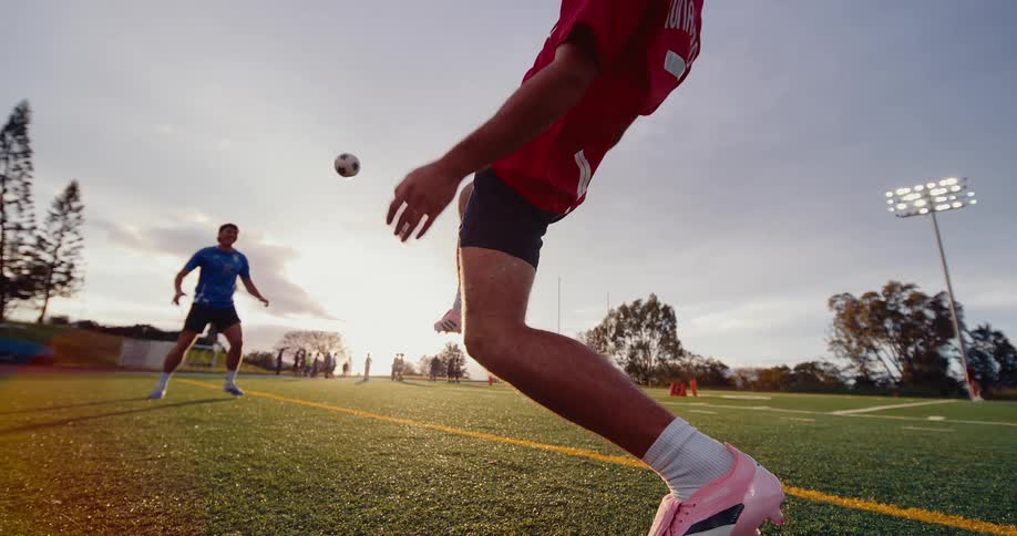 A man wearing a red sports jersey and pink shoes uses his feet to keep a soccer ball in the air while another man in a blue shirt stands on a green field during a sunset