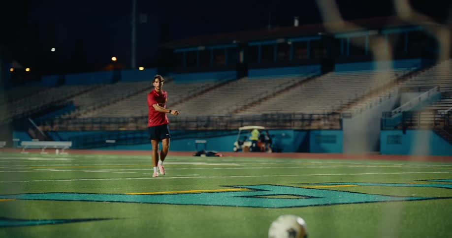 Player kicking a soccer ball on a sports field seen through the goal net