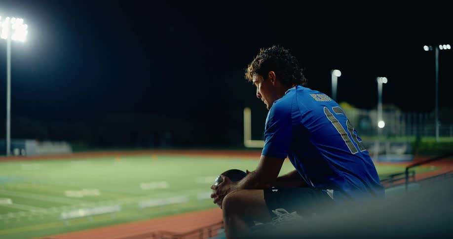 A male athlete sitting on a bench inside a stadium watching the empty pitch at night