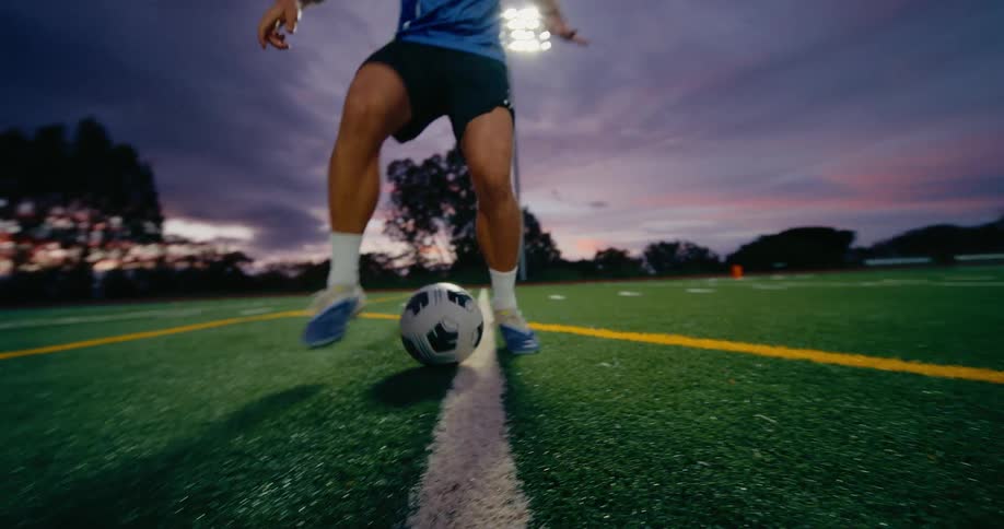 A soccer player in a blue kit dribbles a ball directly toward a low camera on a green turf field at dusk while stadium lights glow against a cloudy purple sky