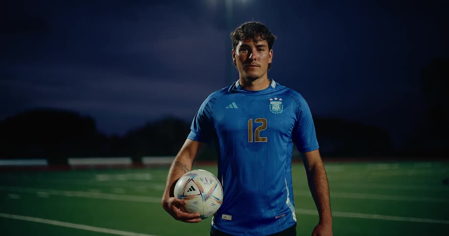 A soccer player stands on a green field at night holding a ball under his arm while wearing a blue argentina jersey with the number 12 the camera pans up from his feet to his face as stadium lights shine brightly in the background