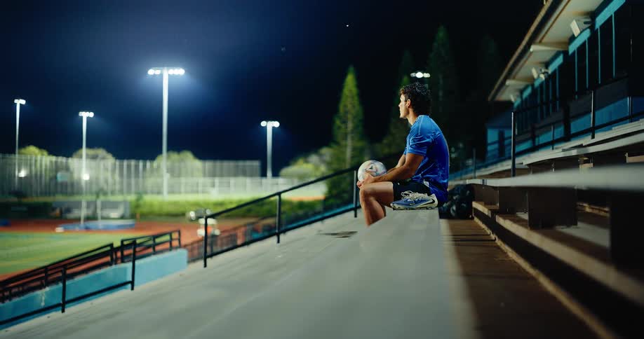 A man with dark curly hair wearing a blue sports jersey sits on outdoor stadium bleachers at night holding a soccer ball and then stands up to walk away while bright field lights shine in the background
