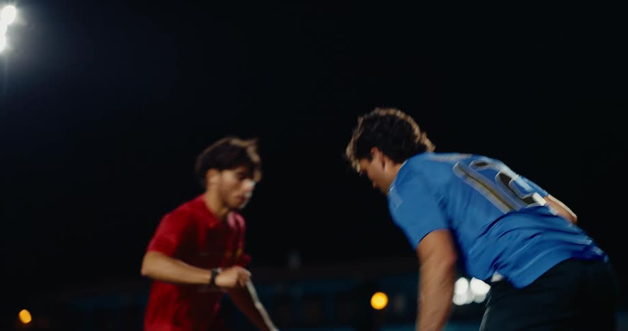 A soccer player in a red jersey skillfully dribbles the ball down the field during a night game under bright stadium lights he successfully maneuvers past a defender in a blue jersey who attempts to slide in and steal the ball