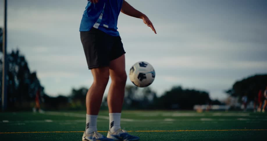 A soccer player wearing a blue jersey with the number twelve juggles a ball with his feet and knees on a green turf field under stadium lights during an evening practice session as the sun sets in the background