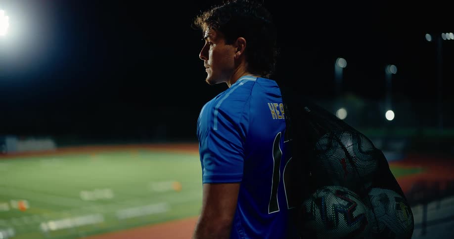 A man walks unto the soccer field with a bag of soccer balls on his back at night