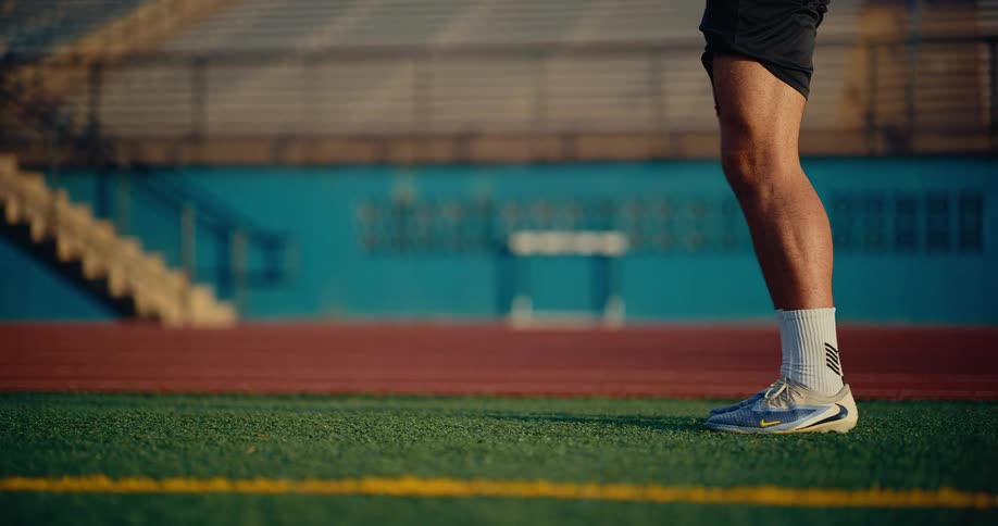 A curly haired man wearing a blue number twelve jersey and black shorts juggles a soccer ball with his feet on a turf field inside an empty stadium during the day