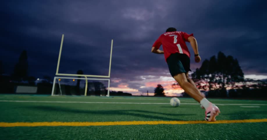 A lowangle view captures a soccer player sprinting dribbling and kicking a soccer ball across the field at dusk his cleats gripping the turf as the goal stands ahead beneath a darkening sky