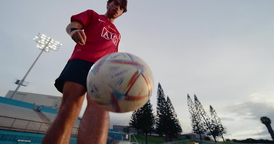 A young man wearing a red jersey and pink cleats juggles a soccer ball on a green field at a stadium
