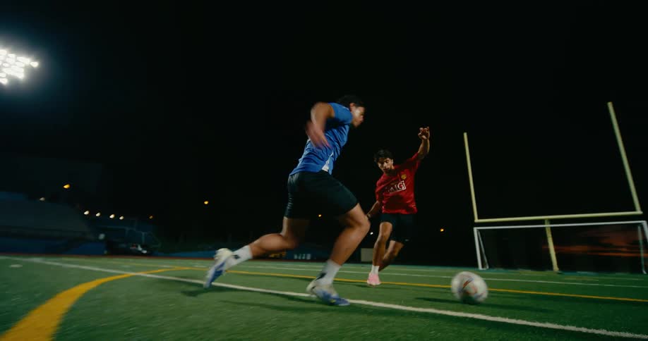 A soccer player in a blue shirt dribbles a ball past a defender in a red jersey on a turf field at night under bright stadium lights near a goal post