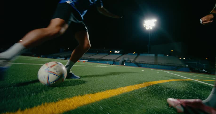 Two players are competing for a ball on a brightly lit sports field at night with the one in blue scoring a goal