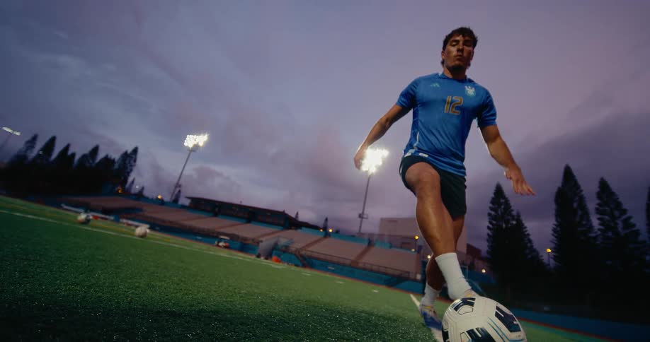 A soccer player in a blue jersey practices footwork drills on a turf field during twilight the camera uses a lowangle perspective to follow his feet as he dribbles the ball along a white boundary line