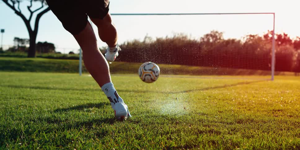 A soccer player kicks a ball into a goal on a grass field during the late afternoon