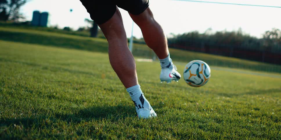 A close view shows the legs and feet of a person wearing white socks and cleats as they run toward and kick a soccer ball on a green grass field during the day