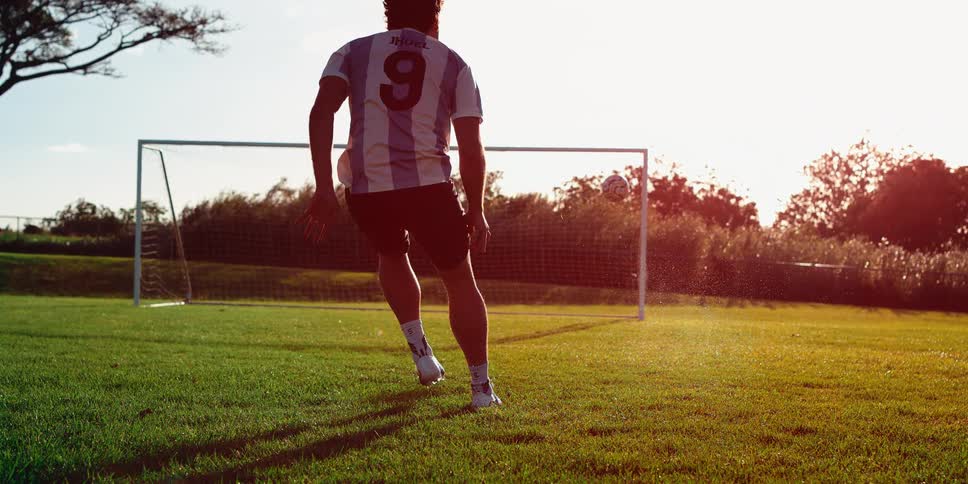 Young adult man kicking a soccer ball into a goal and then jumping on the air on a field surrounded by vegetation outdoors
