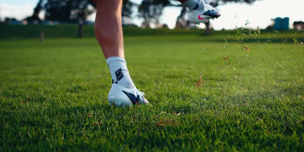 A person wearing white soccer cleats and socks kicks a white and gold soccer ball on a green field the impact sends the ball flying forward and kicks up a spray of grass and dirt from the turf