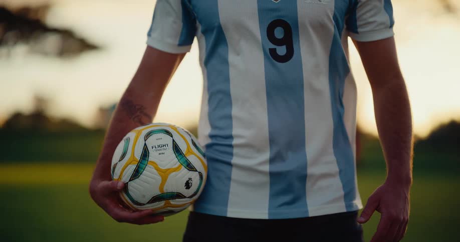 A close view shows the midsection of a man wearing a light blue and white striped sports jersey with the number nine while he holds a soccer ball in his hands on a field during a sunset