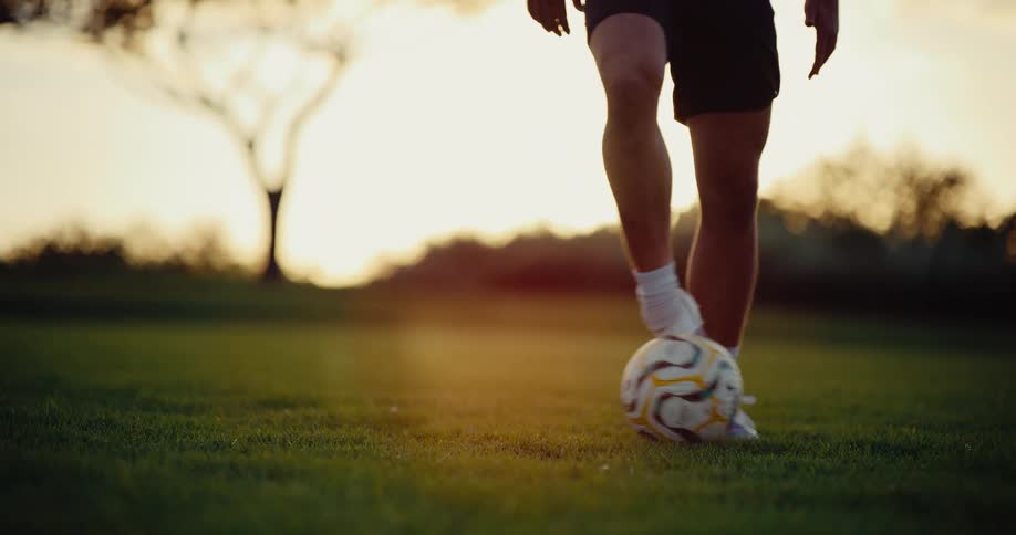 A soccer player practices footwork by rapidly tapping and rolling a ball between their feet on a grass field the scene is captured during sunset casting a warm golden light over the player and the grassy terrain