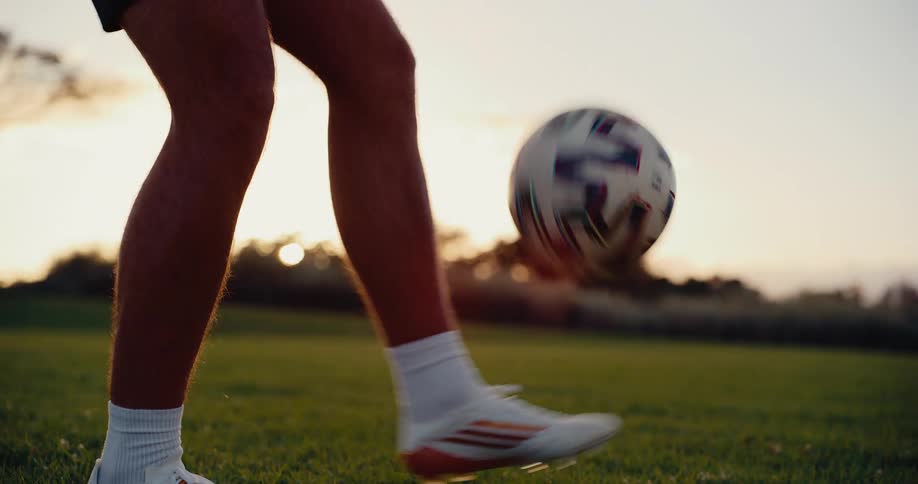 A man in white cleats and shorts juggles a soccer ball on a green grass field during a golden hour sunset