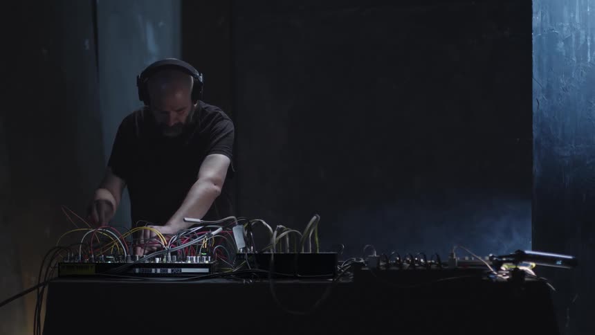 A man with a beard and headphones stands in a dark moody studio focused on manipulating a complex modular synthesizerbright cables snake across the table as he adjusts knobs and patch points his movements highlighted by a single side light source