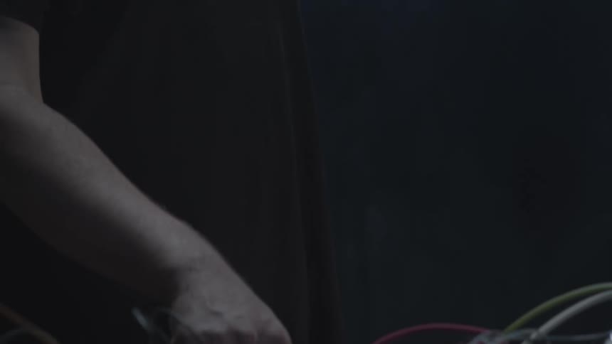 A bearded man in a dark shirt gazes downward with a focused expression as he works in a dimly lit studio his hand reaches out to manipulate a dense network of colorful patch cables and knobs on a modular synthesizer