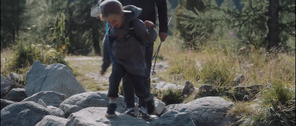 Boy wearing a baseball cap jumping between rocks with an adult walking behind him and holding a safety harness that is strapped on his body next to a grove outdoors during day time
