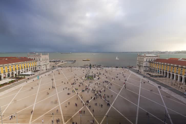 A highangle timelapse shows a large public square with a geometric paved pattern filled with moving crowds and framed by yellow buildings in the background dark clouds loom over a wide body of water where several boats are visible