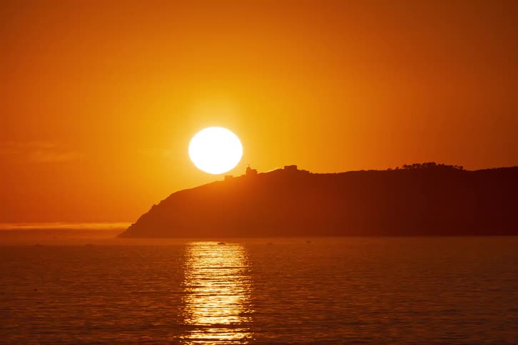 This timelapse video captures a bright sun slowly descending behind a dark silhouetted hillside overlooking a calm sea as the sun disappears the vibrant orange glow across the sky and the shimmering reflection on the water gradually fade into a deeper