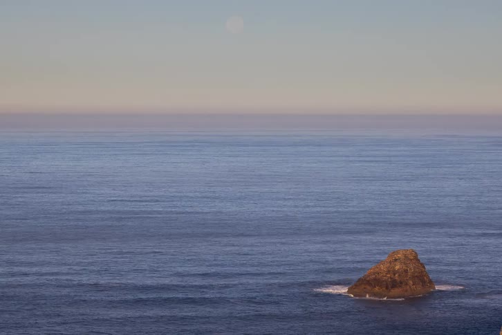 A wide shot of a calm blue ocean with a single small rocky island protruding from the waterthe horizon features a soft gradient of light orange and grey with a faint pale moon visible in the upper center of the frame