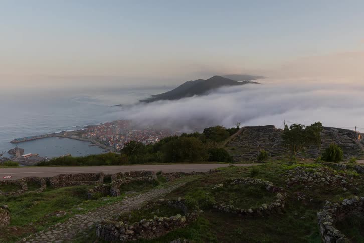 Low lying clouds pass over a coastal city as people walk on a trail and road near low stone walls