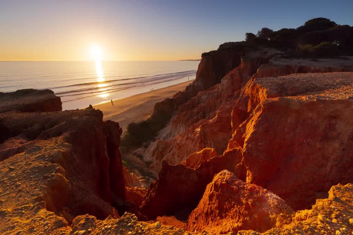 Sun setting on the horizon over the ocean beside rocky cliffs illuminated by golden light