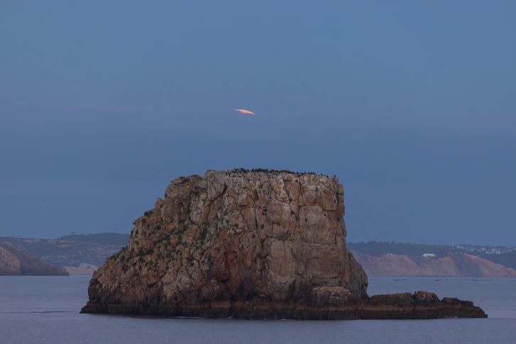 A large rock formation in the ocean under a purple twilight skyas the sky darkens a bright yellow full moon rises from behind the rock and ascends into the night