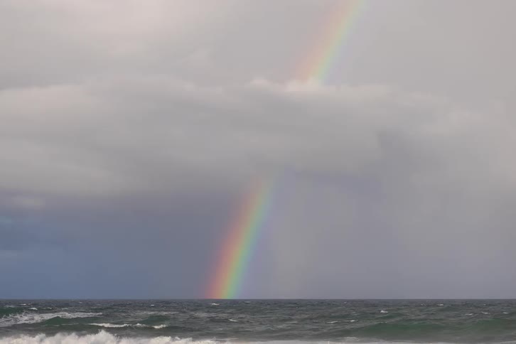 A bright rainbow emerges from a dark horizon where turbulent ocean waves crash under a heavy gray sky