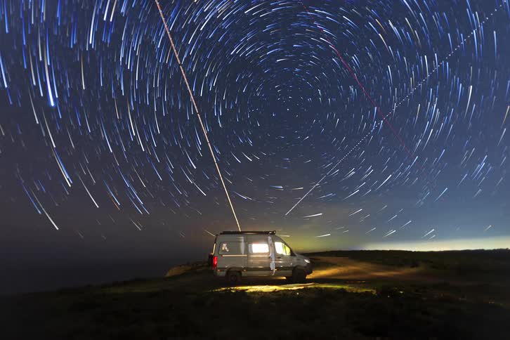 A camper van sits parked on a grassy cliff at night under a revolving sky of circular star trails while its interior and exterior lights illuminate the immediate ground and a distant horizon glows