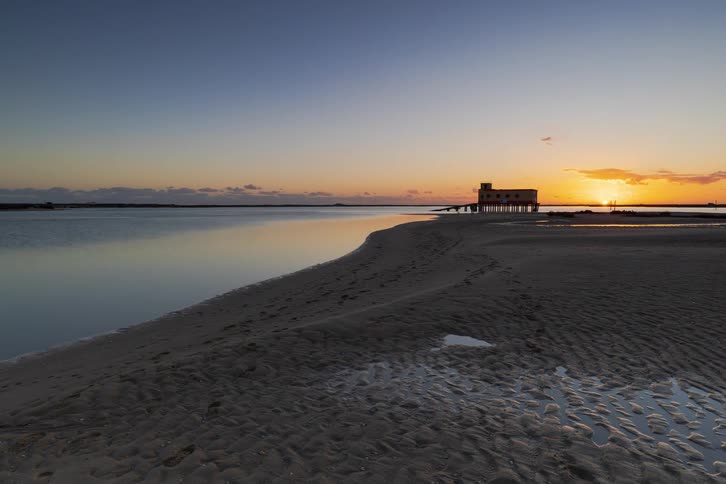 A wide sunset landscape shows a coastal beach with wet rippled sand and calm water as a small dog and people walk toward a silhouetted building on the horizon under a clear orange and blue sky