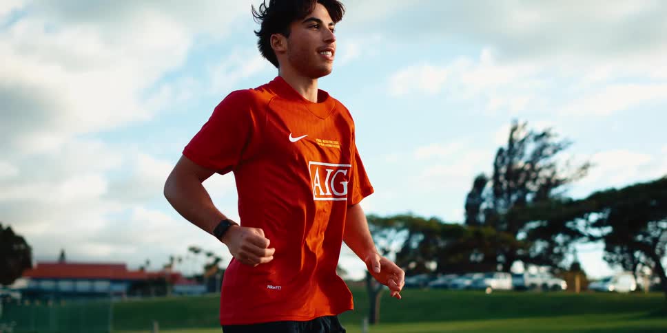 Young man running in an open field wearing a red sports shirt under a partly cloudy sky