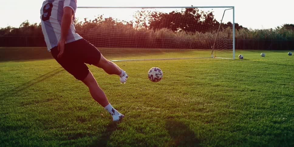 A soccer player kicking a ball toward a goal on a grassy field at sunset