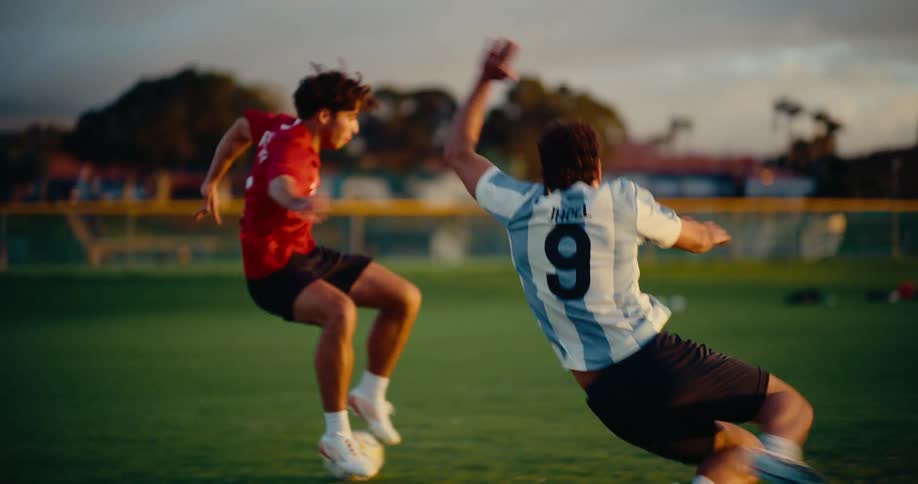 Two soccer players run across the field following the ball and one player falls on the ground