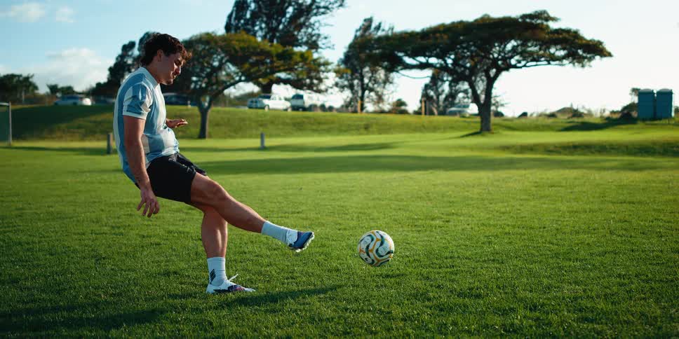 A soccer player wearing a light blue and white striped jersey kicks a ball across a sunny green fieldthe camera follows the balls movement as the player completes his followthrough under a clear sky