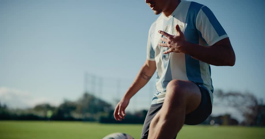 A young man wearing a striped jersey and black shorts practices juggling a soccer ball with his knees and feet on a bright green grass field under a clear blue sky during a daytime training session at an outdoor sports complex