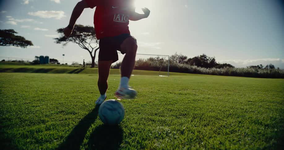 A soccer player in a red jersey performs a skill move while dribbling a ball on a green grass field during sunset with a goal and trees in the background
