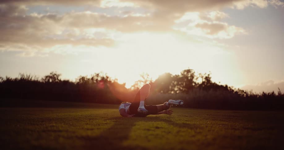 A soccer player performs an acrobatic bicycle kick on a grassy field during a golden sunsetafter the strike he lands on his back and sits up while the sun flares directly behind him