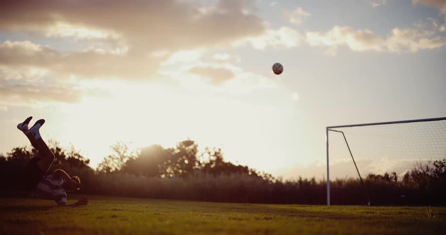 Young adult soccer player bycicle kicking a soccer ball in front of a goal and missing it on a field with vegetation in the backgound outdoors during dusk or dawn