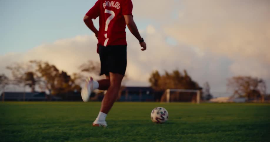 A male soccer player in a red jersey dribbles a ball across a green grass field during golden hour while goal posts and trees sit in the distance under a cloudy sky
