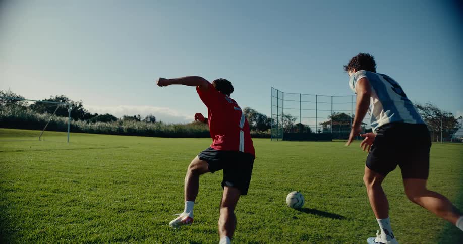 Two young men are playing soccer on a grassy field under a clear blue sky the player in the blue and white striped jersey dribbles the ball past the player in the red shirt as they run across the field
