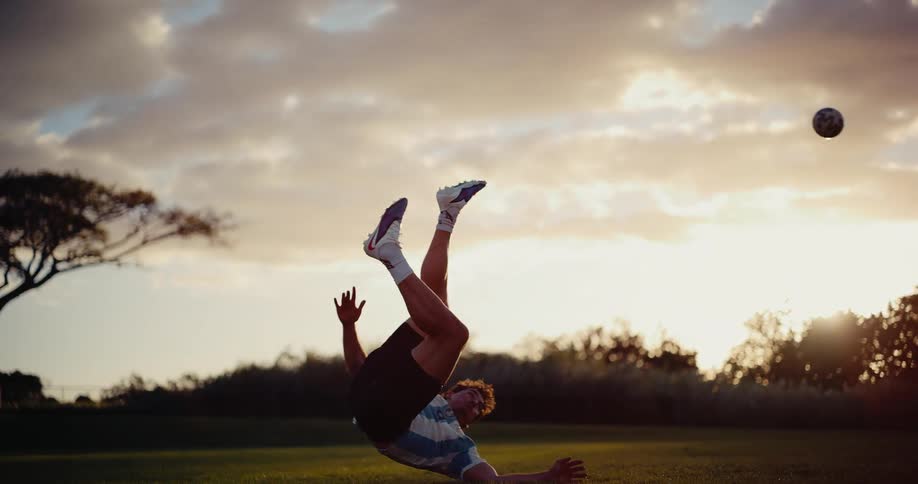 A soccer player in an argentina jersey attempts a dramatic bicycle kick during a golden hour sunset he makes contact with the ball midair before falling onto the grass as the sun glows in the background
