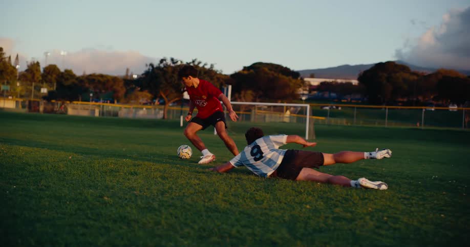 Two soccer players run across the field following the ball and one player falls on the ground and the other player takes the ball